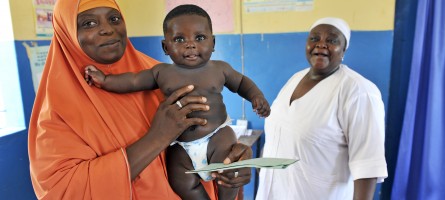 A mother in Nigeria holds her infant; she is in a clinical setting. A nurse smiles in the background.